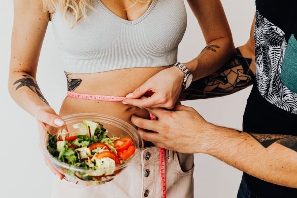 A couple measuring waistline with a tape and holding a fresh salad bowl, focusing on fitness and weight loss.