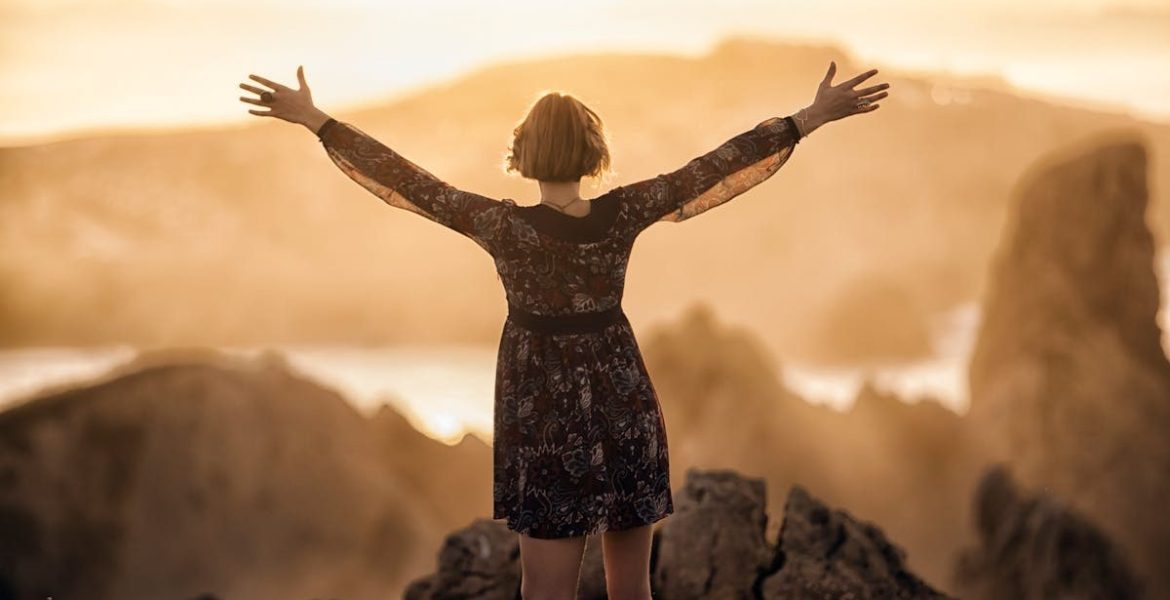 A woman stands with open arms on a mountain at sunset, embracing nature's beauty.