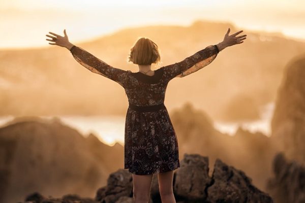 A woman stands with open arms on a mountain at sunset, embracing nature's beauty.